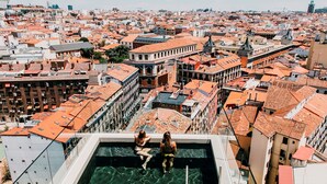 A rooftop pool with two people sitting at the edge, overlooking a densely packed urban area with red-tiled roofs. - Dear Hotel Madrid (Madrid)
