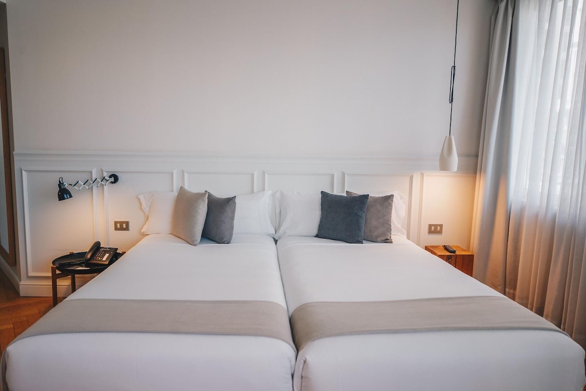 A double bed with white bedding and two pillows, a bedside table with a lamp, and a phone.