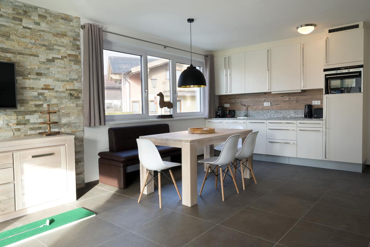 A modern kitchen with a dining area, stone wall backsplash, and a large window with curtains.