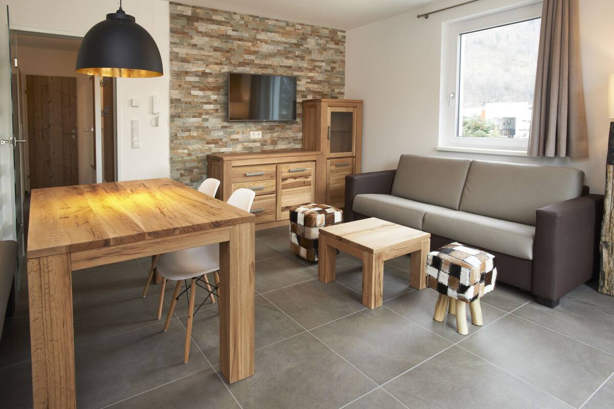 A modern living room with a wooden dining table, a sofa, and a television mounted on a brick wall.