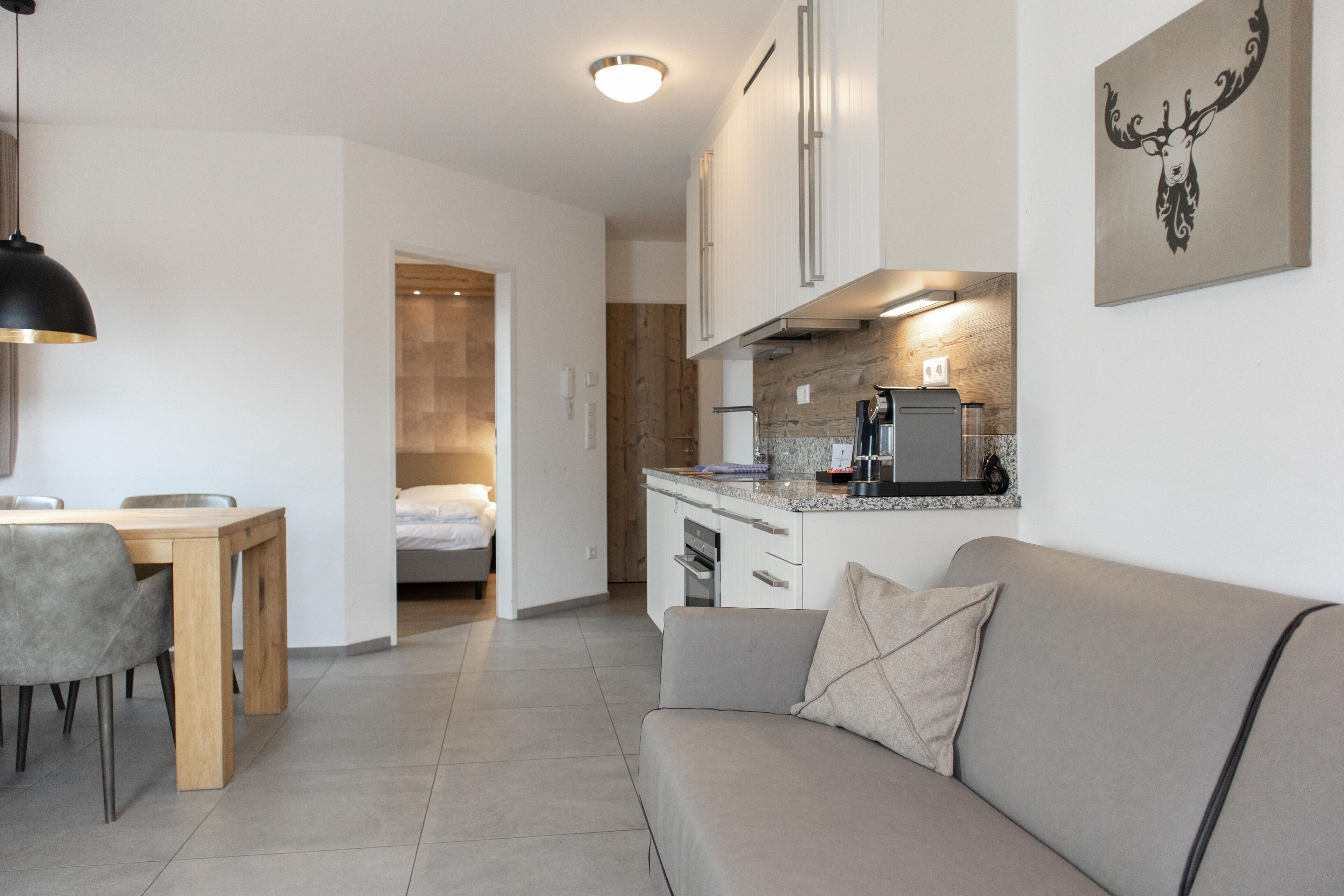 A modern kitchen with a built-in oven and a coffee machine, a dining area with a wooden table and chairs, and a living area with a sofa and a painting on the wall.