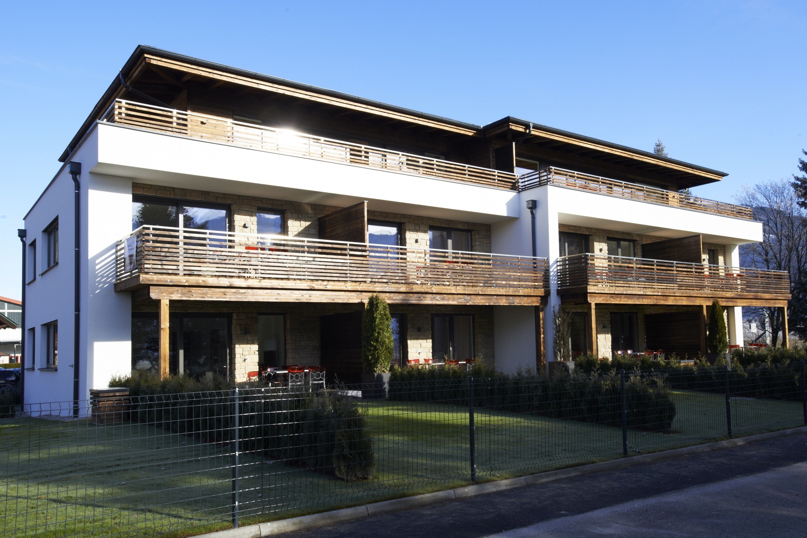 A modern two-story building with a balcony on the upper floor, a covered entrance, and a fenced front yard.
