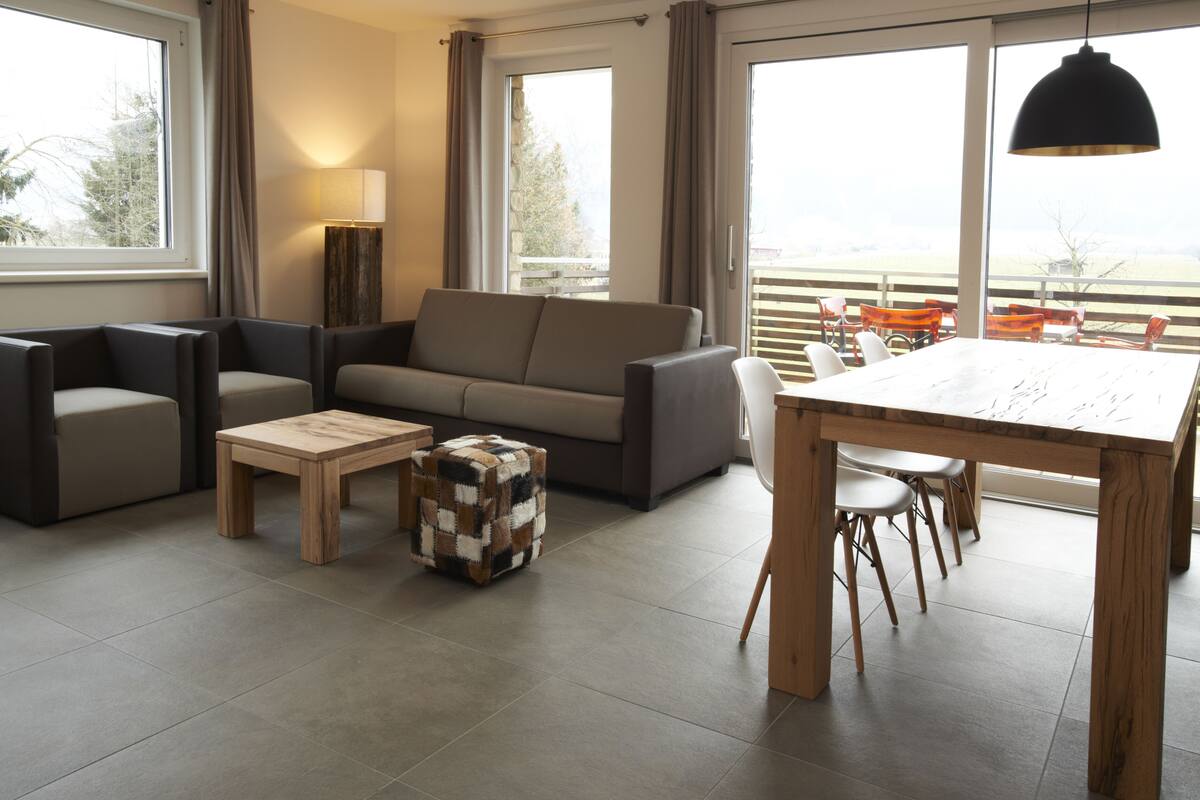 A modern living room with a sofa, coffee table, and dining table. There are large windows providing a view of the outdoors.