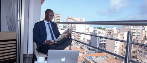 A man sitting on a balcony with a laptop and a cup, overlooking a cityscape.