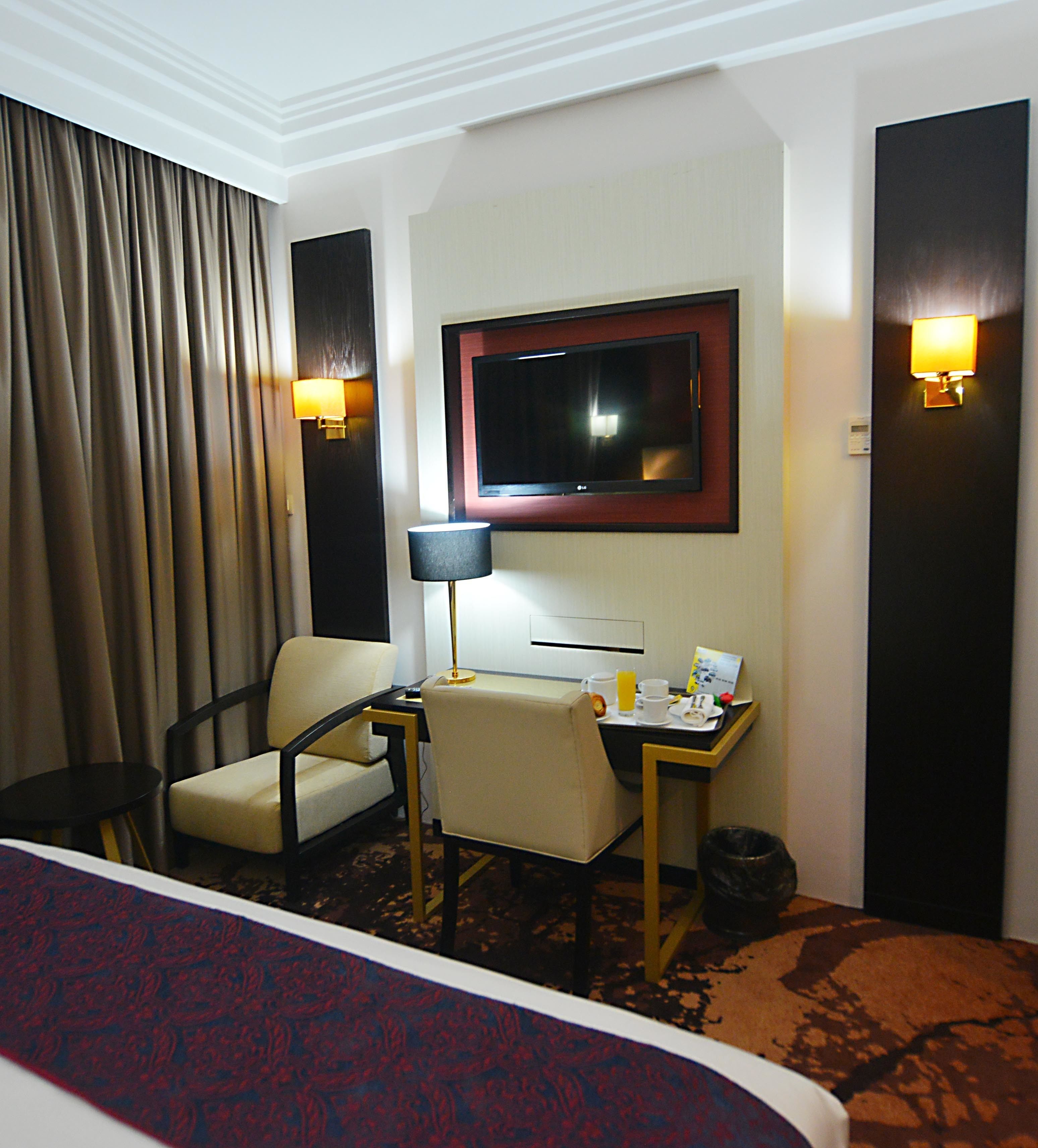 A hotel room with a bed, a desk with a lamp, a chair, a TV mounted on the wall, and a trash bin.