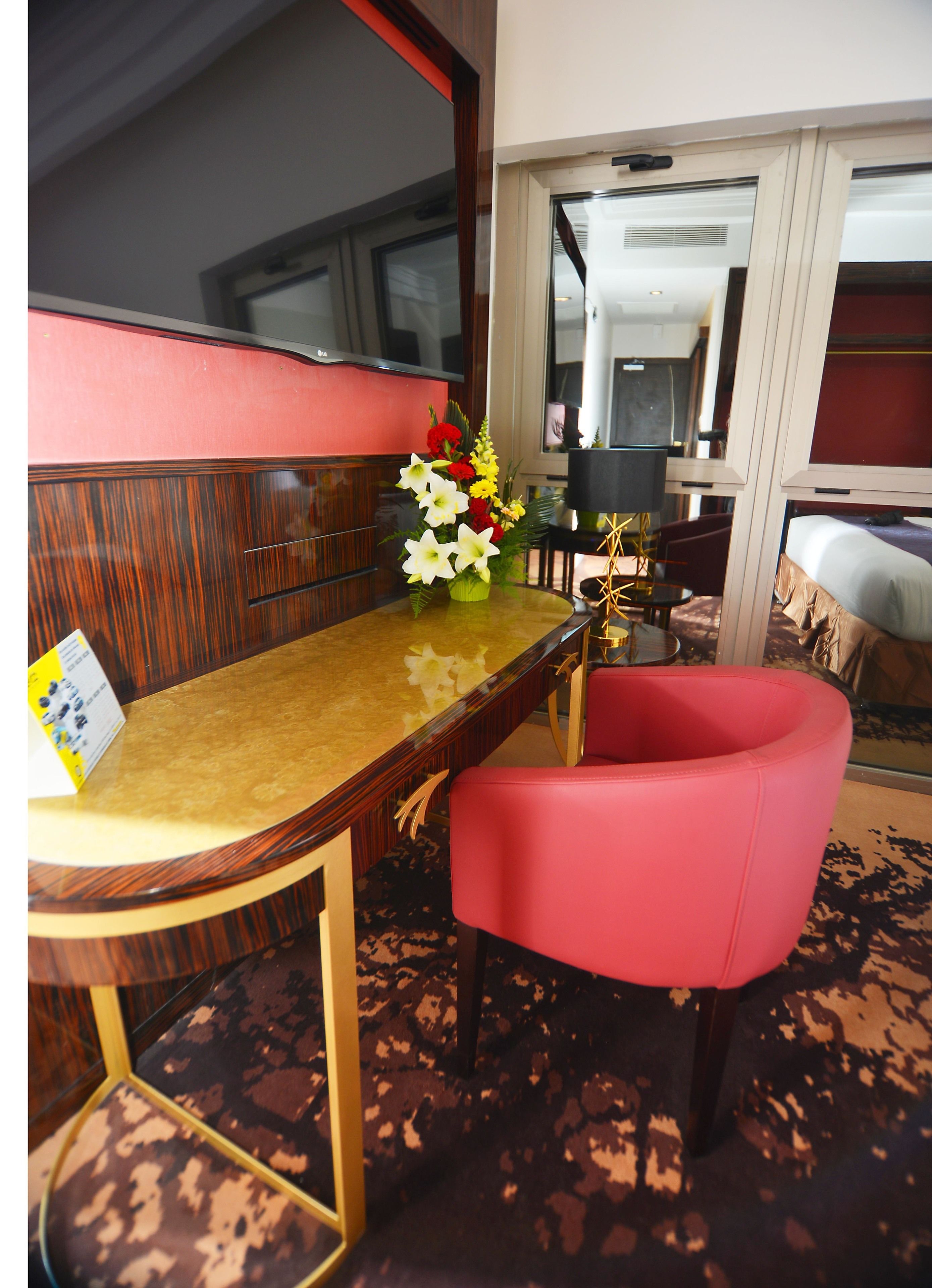 Habitación de hotel con un mostrador de recepción de madera, una silla roja, una mesita con un jarrón de flores y una cama con cabecera roja.