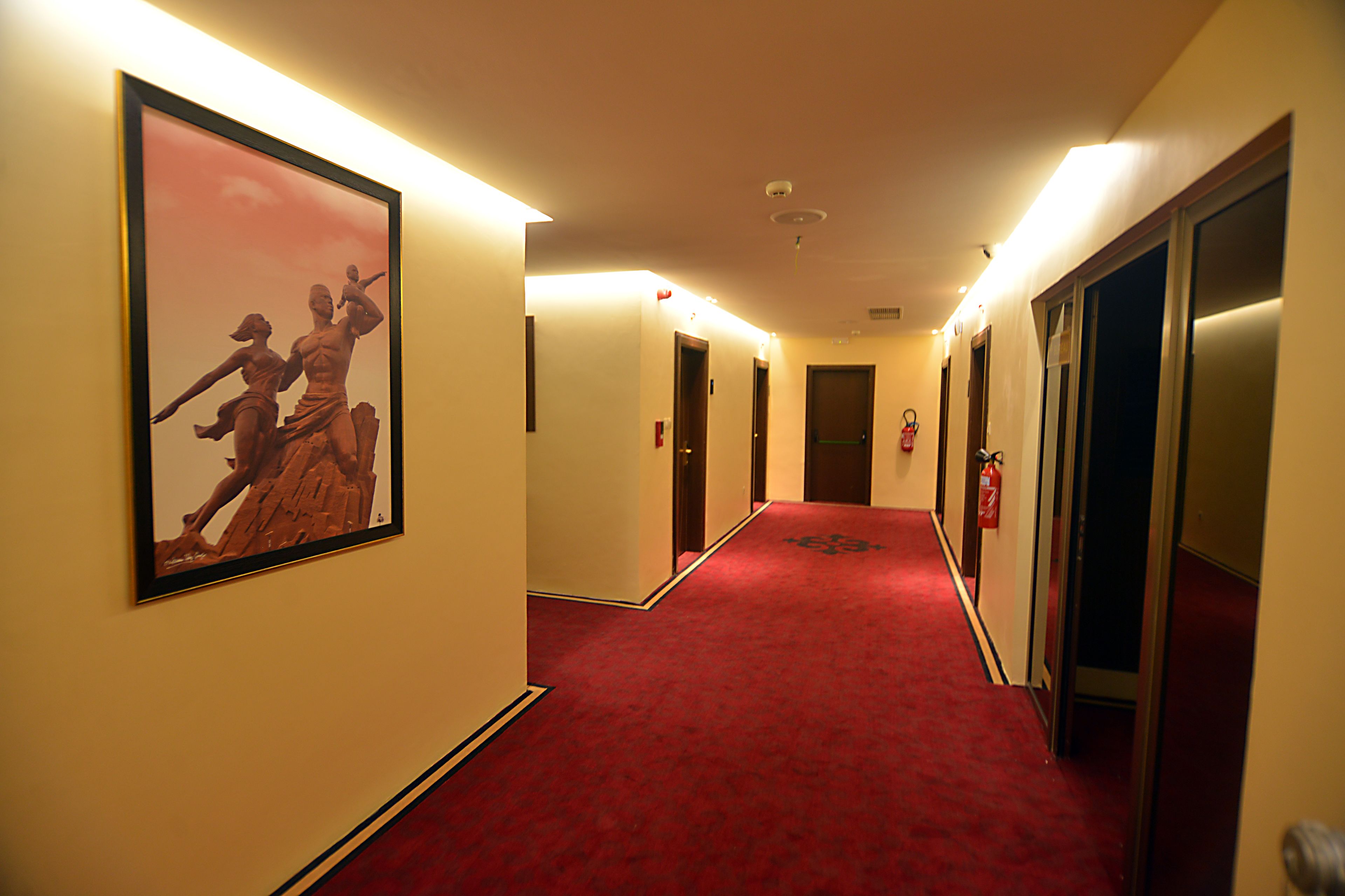 A hotel hallway with a red carpet, framed artwork on the walls, and doors leading to rooms.
