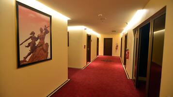 A hotel hallway with a red carpet, framed artwork on the walls, and doors leading to rooms.