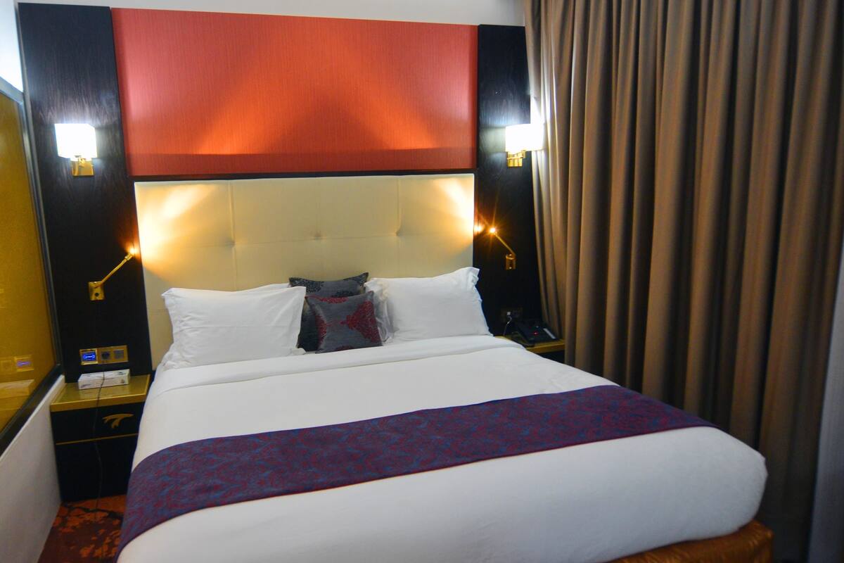 Une chambre d’hôtel avec un grand lit, une tête de lit dotée d’un éclairage intégré, une table de chevet avec un téléphone et un rideau.