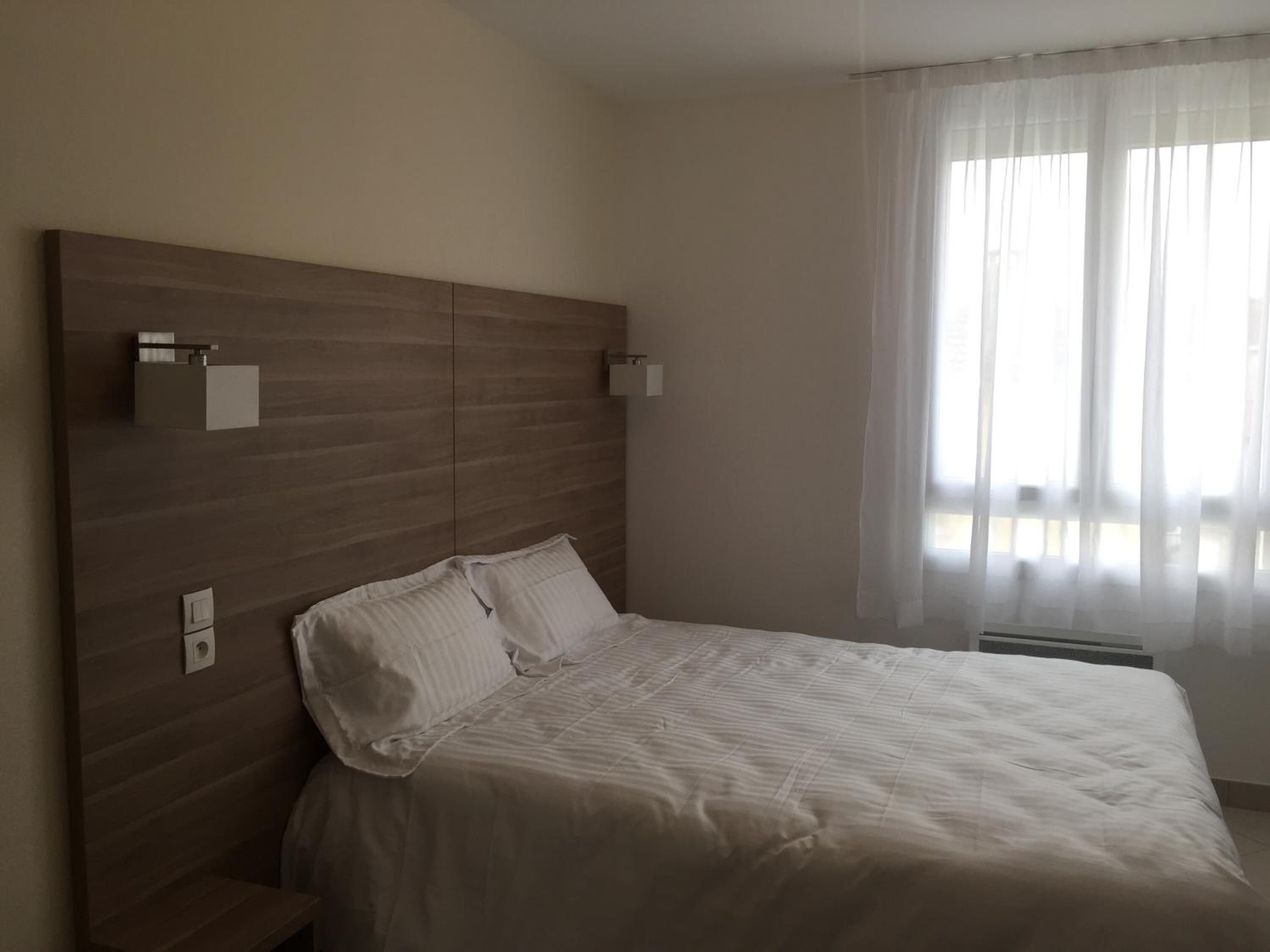 A bedroom with a wooden headboard, two bedside tables, a bed with white bedding, and a window with sheer curtains.