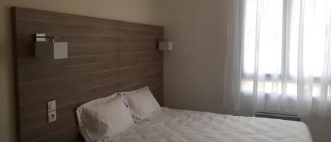 A bedroom with a wooden headboard, two bedside tables, a bed with white bedding, and a window with sheer curtains.