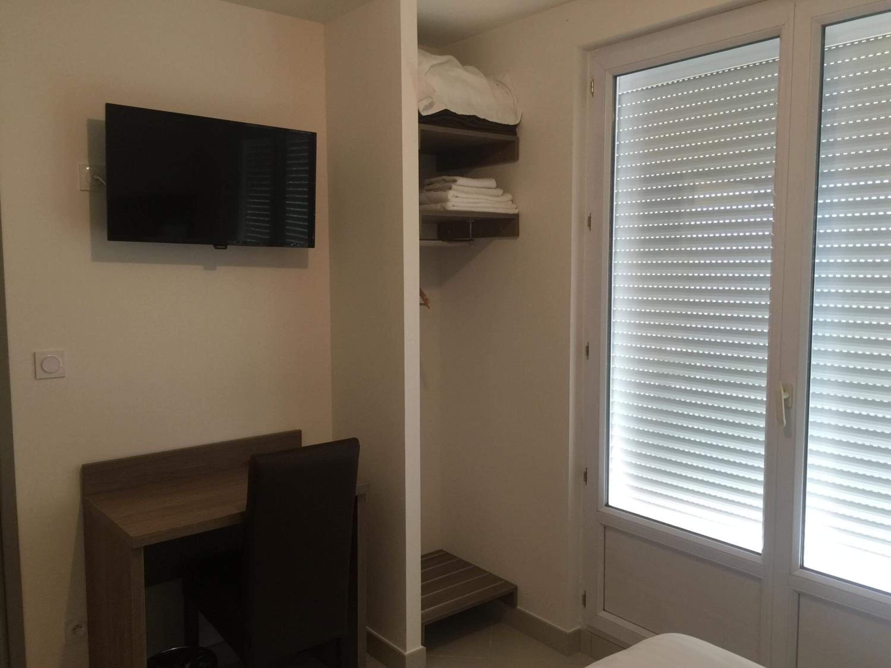 A small, well-lit room with a desk, chair, and a television mounted on the wall. There is a sliding door with blinds, and a shelf with folded towels.