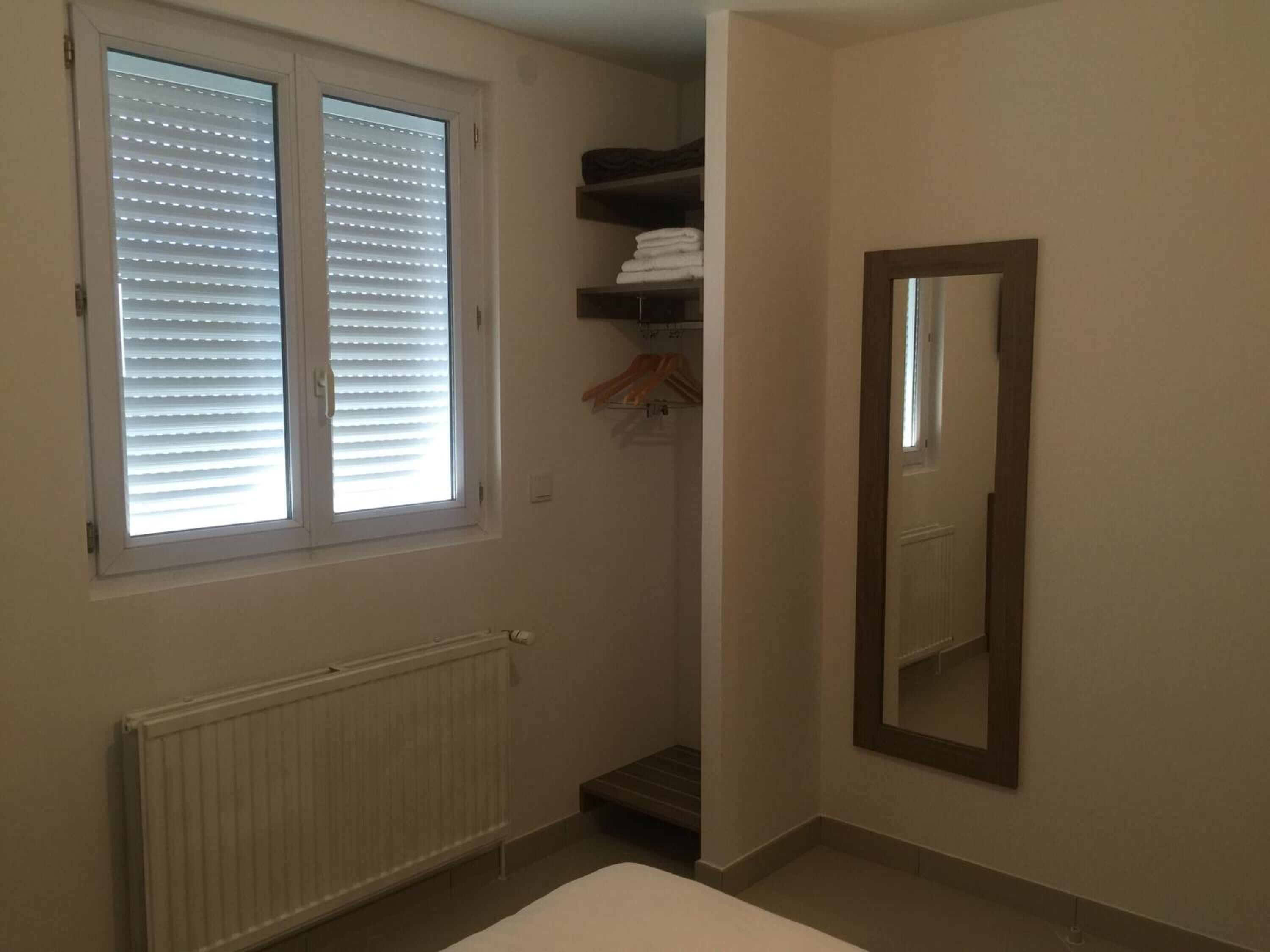 A small, well-lit room with a radiator, a window with blinds, a mirror, and a shelf with folded towels.