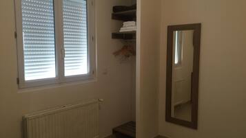Una habitación pequeña y luminosa, con un radiador, una ventana con persianas, un espejo y un estante con toallas dobladas.