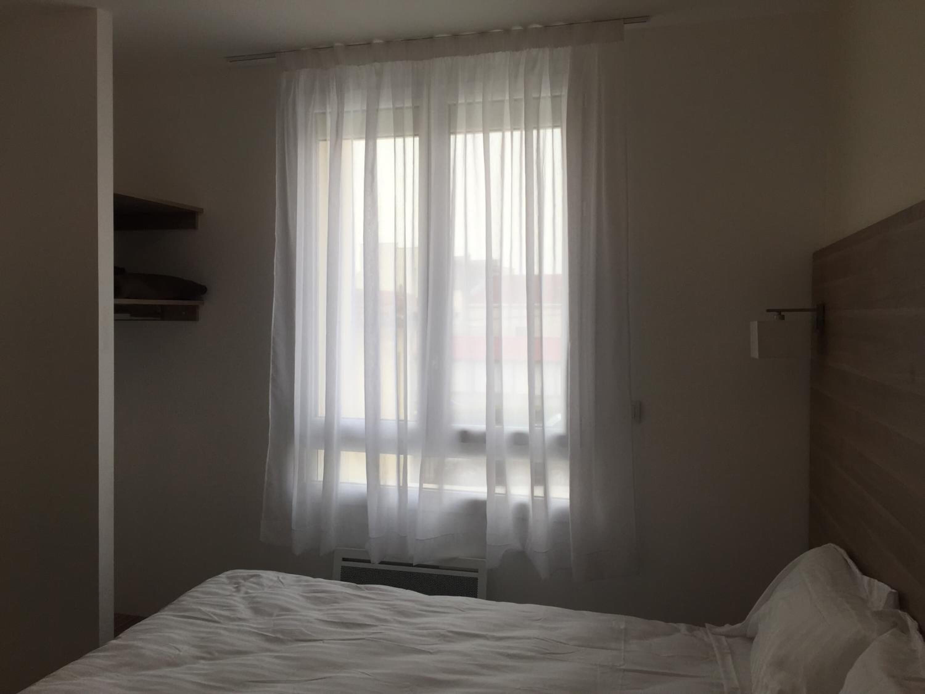 Une chambre à coucher avec un lit, une fenêtre avec des rideaux légers, un radiateur et une étagère fixée au mur.