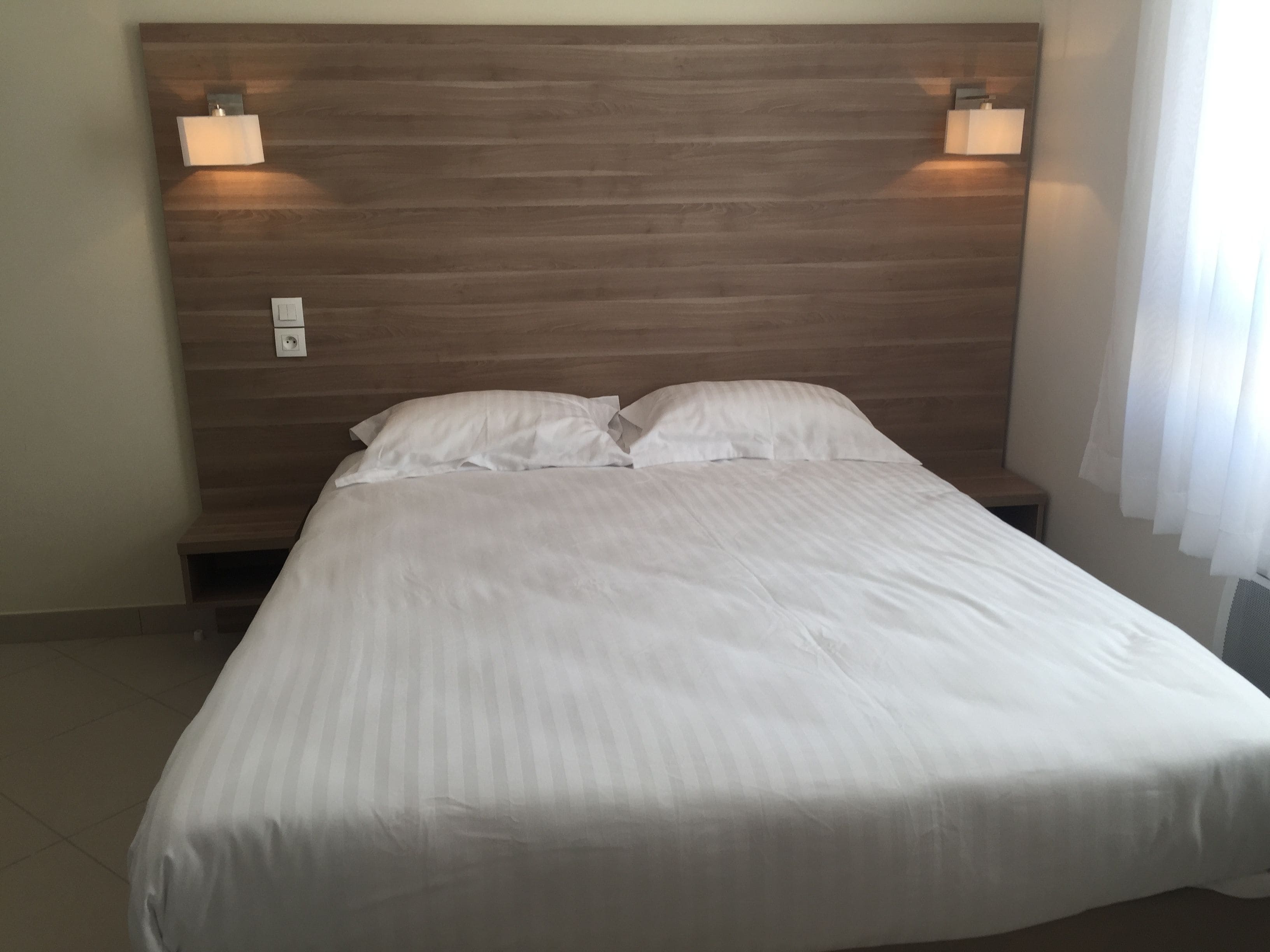 Una cama bien hecha con sábanas blancas, cabecera de madera y dos lámparas fijadas a la pared.