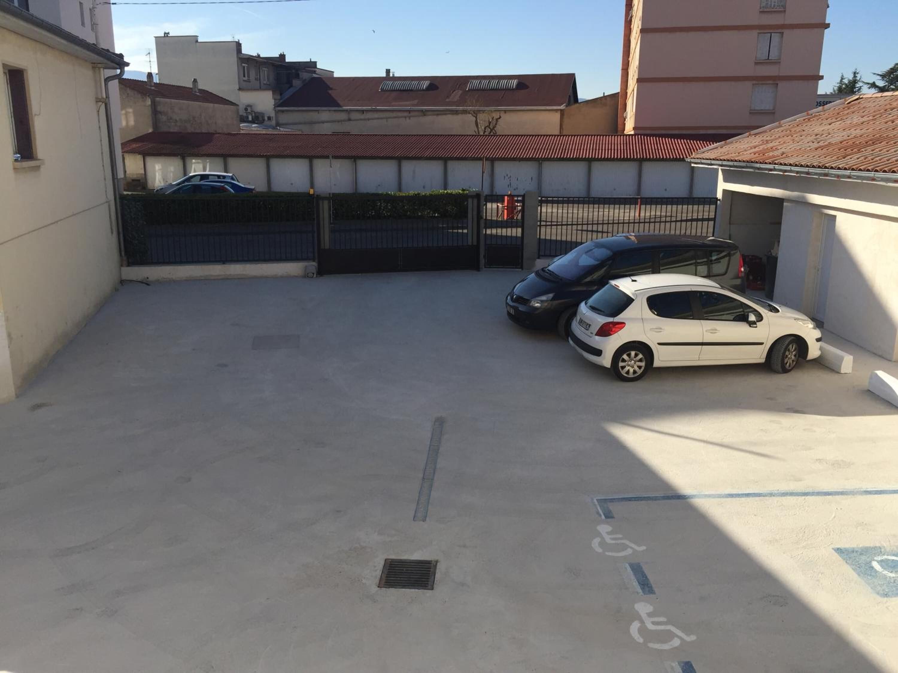 Un espace de stationnement avec deux voitures, un symbole indiquant l’accessibilité aux personnes à mobilité réduite et, au fond, un bâtiment avec un toit rouge.