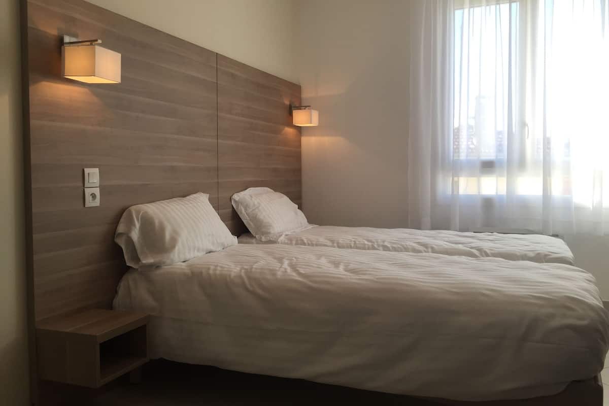 A bedroom with two beds, wooden headboard, bedside lamps, and a window with sheer curtains.