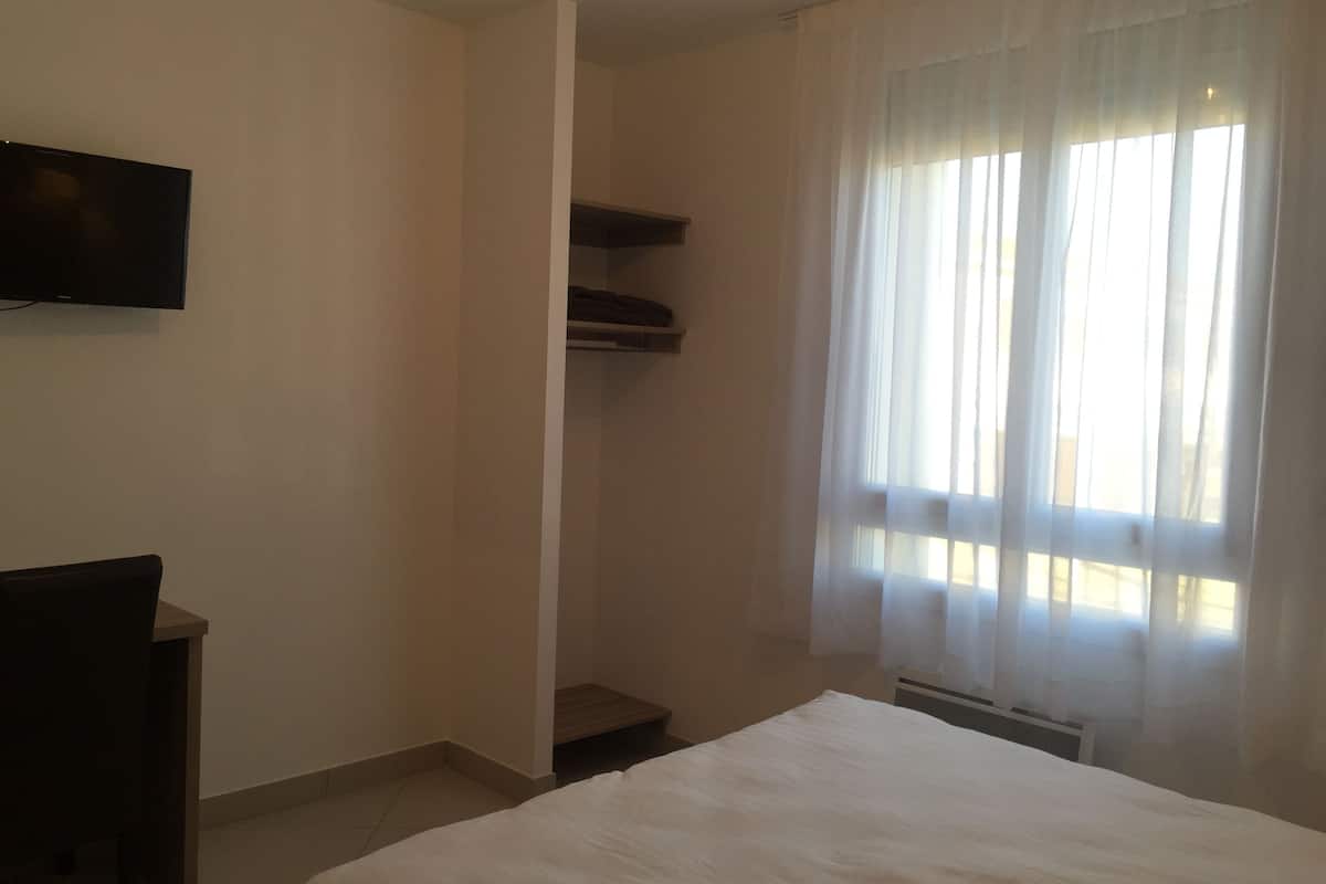A bedroom with a bed, a TV mounted on the wall, a chair, and a window with sheer curtains.