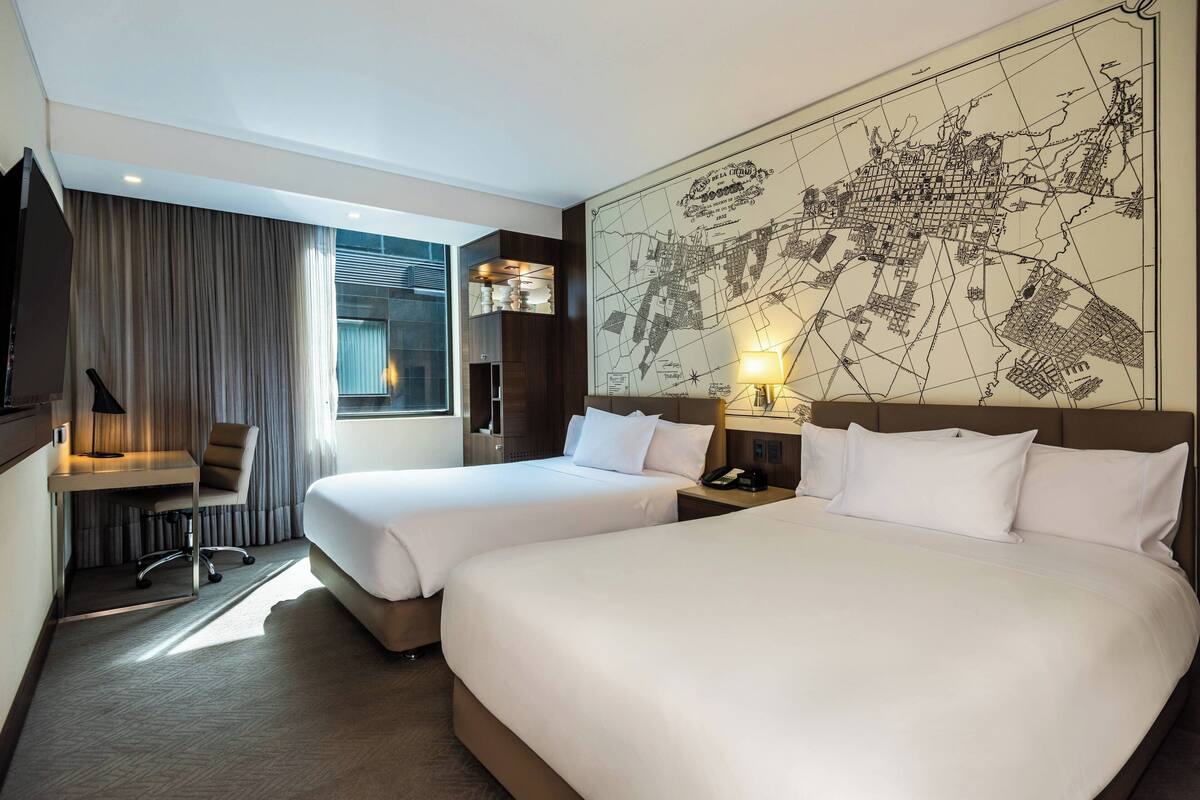 Une chambre d’hôtel avec deux lits, un bureau, une chaise, une carte accrochée au mur et une fenêtre avec des rideaux.