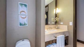 Un baño con inodoro, lavamanos y espejo. Hay dos cuadros enmarcados en la pared, un florero con flores y un perchero con toallas.