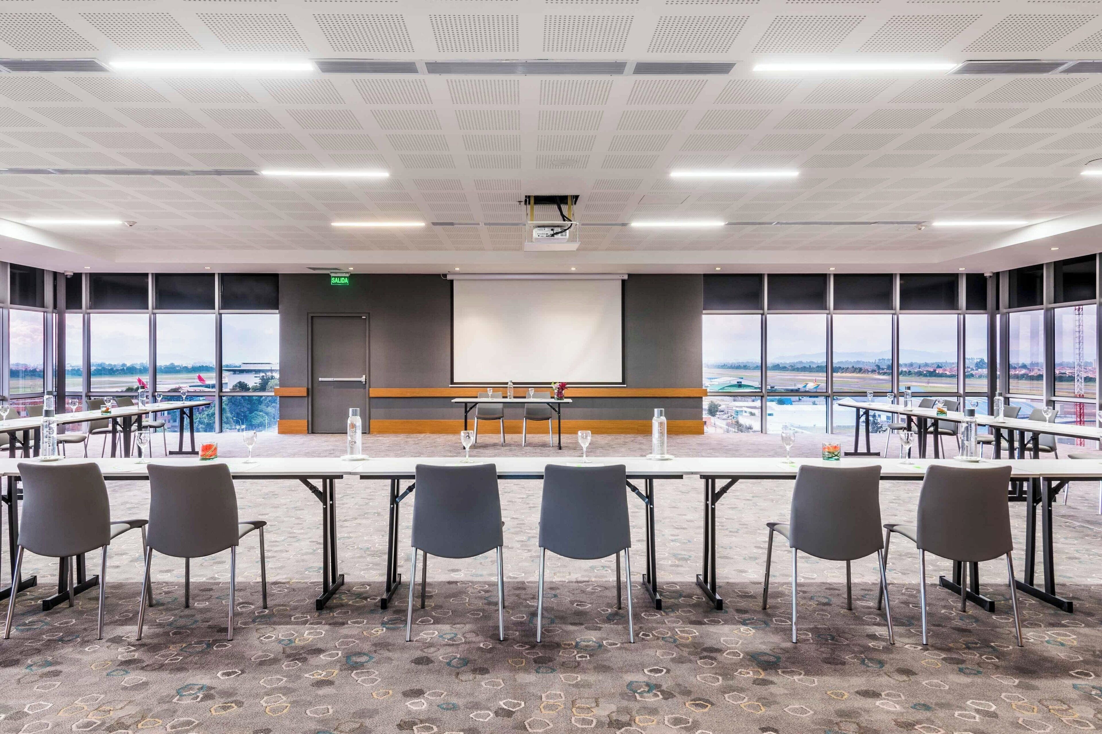 A conference room with a large table, chairs, a projector screen, and large windows offering a view of the cityscape.
