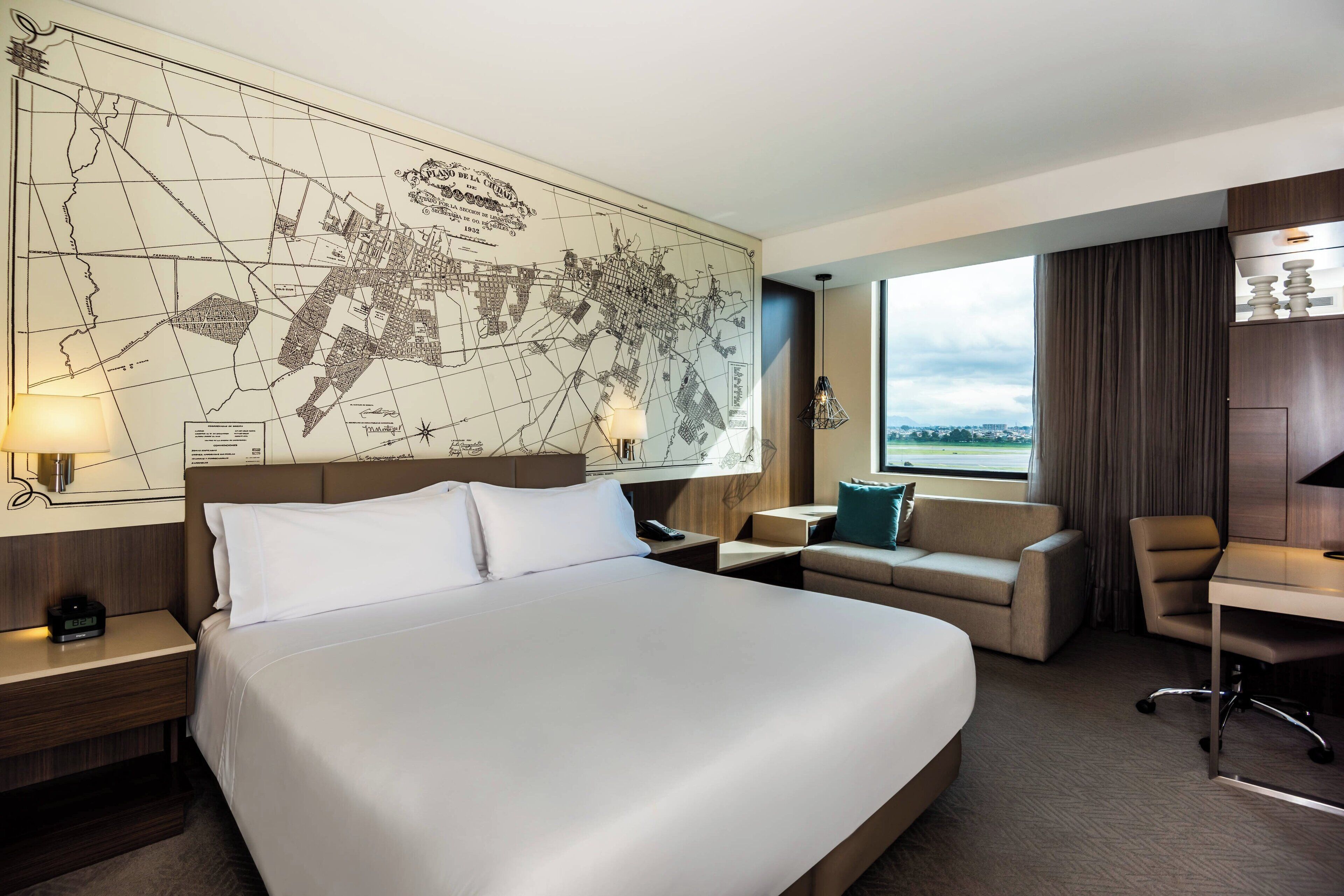 A hotel room with a large bed, a desk, a chair, a sofa, a map on the wall, and a window with a view.