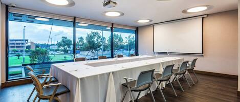 Un salón de conferencias con una larga mesa, sillas, una pantalla de proyector y ventanas grandes que dan a un parque y edificios.