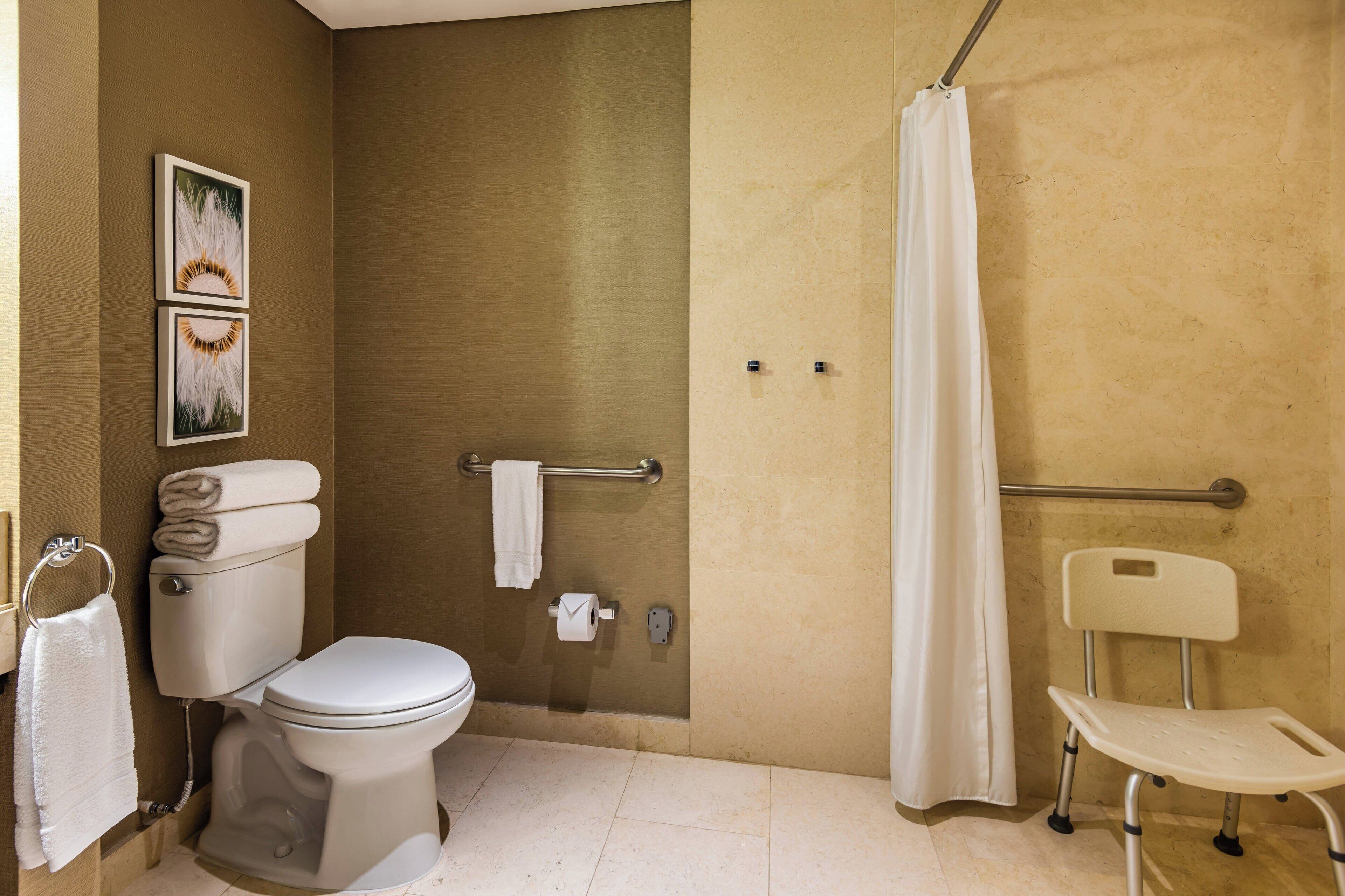 A bathroom with a toilet, a shower curtain, a grab bar, and a wheelchair-accessible chair.