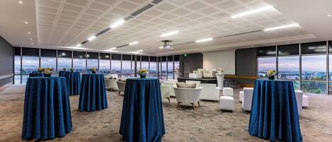 A conference room with multiple tables covered in blue tablecloths, a patterned carpet, and large windows offering a city view.
