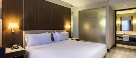 A modern hotel room with a large bed, bedside lamps, a nightstand, a wooden headboard, and a bathroom visible through an open door.