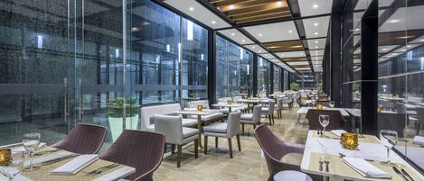 A modern restaurant with glass walls, multiple dining tables, and wicker chairs.