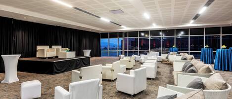 Una amplia sala de conferencias con un escenario, sillas blancas y un gran ventanal con vistas a la ciudad.