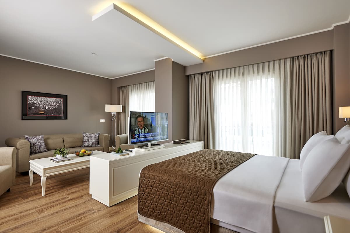 Una habitación de hotel moderna con una cama grande, un sofá, una mesa de centro, un televisor y un ventanal con cortinas.