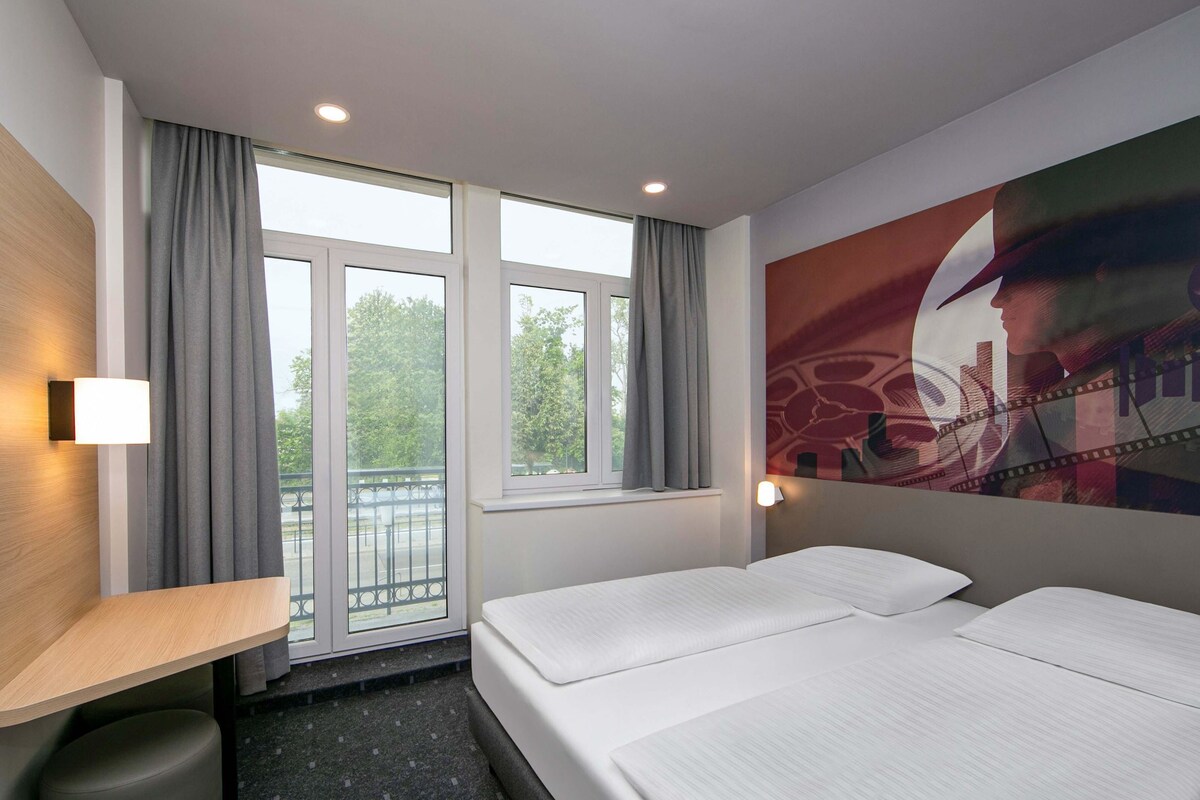 A hotel room with a bed, a desk, a chair, a window with curtains, and a wall-mounted light.