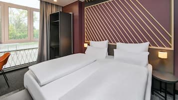 A modern hotel room with a large bed, a desk, and a chair. There is a window with a view of trees and a balcony.