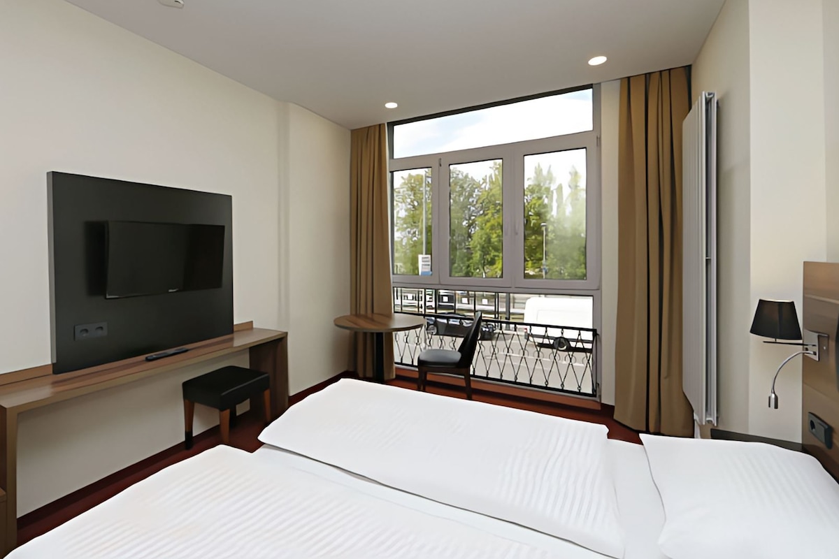 Habitación de hotel con cama, televisor montado en la pared, ventana con vistas al exterior y mesita de noche.
