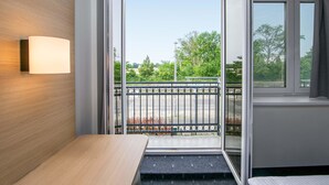 A modern hotel room with a wooden wall, a bedside table, a window with a view of a park, and a balcony with a metal railing. - B&B HOTEL Berlin-Adlershof (Berlin)
