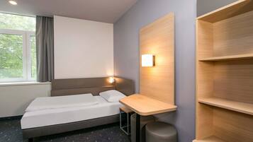 A hotel room with a bed, a small table, a wall-mounted shelf, and a window with curtains.