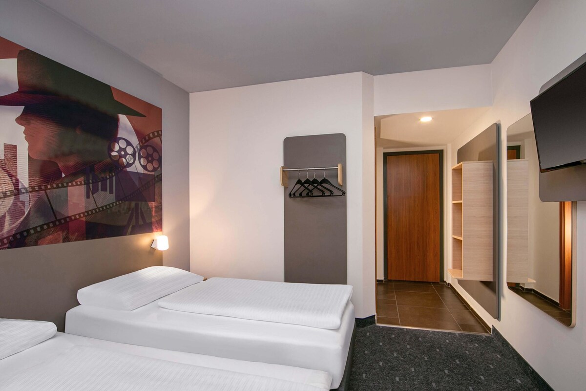 A hotel room with two beds, a wall-mounted TV, a coat rack, and a hallway leading to a bathroom.