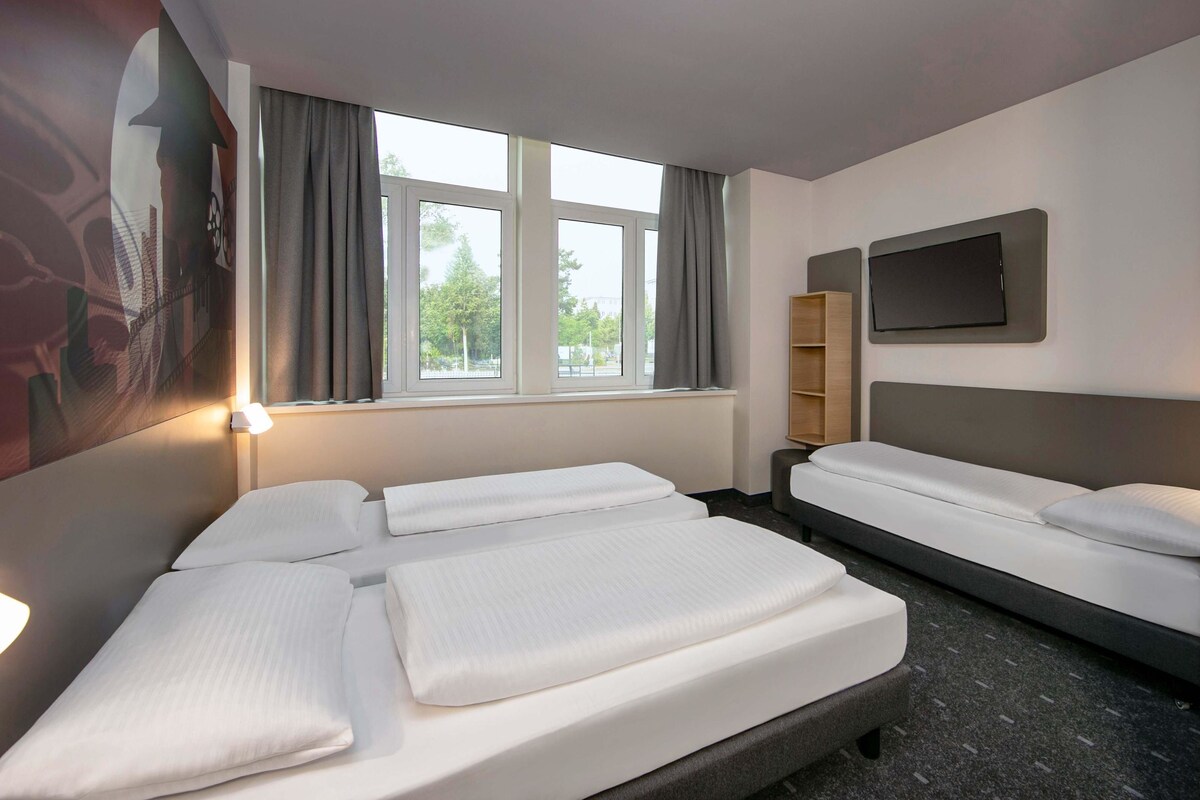 A hotel room with two beds, a TV mounted on the wall, a window with curtains, and a lamp on the left side.