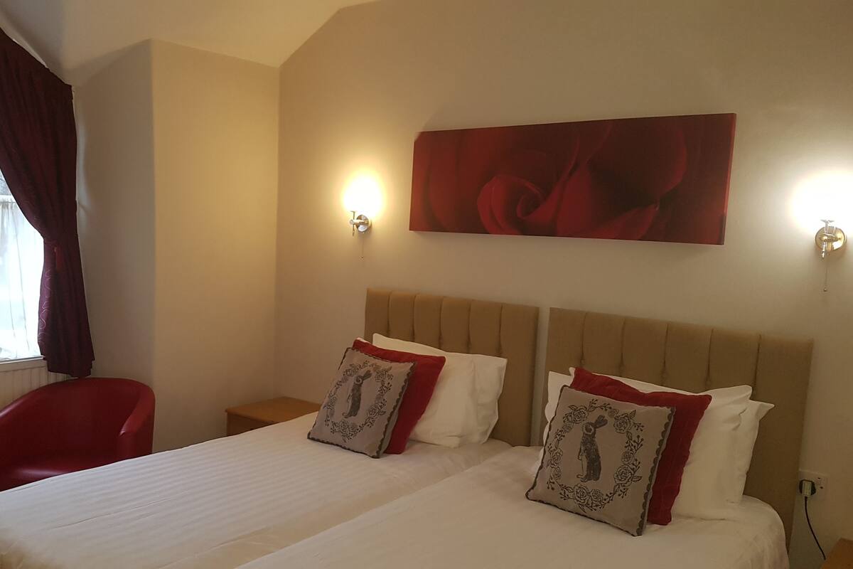 A hotel room with a large bed, two bedside tables, a red chair, a window with curtains, and a wall-mounted artwork.