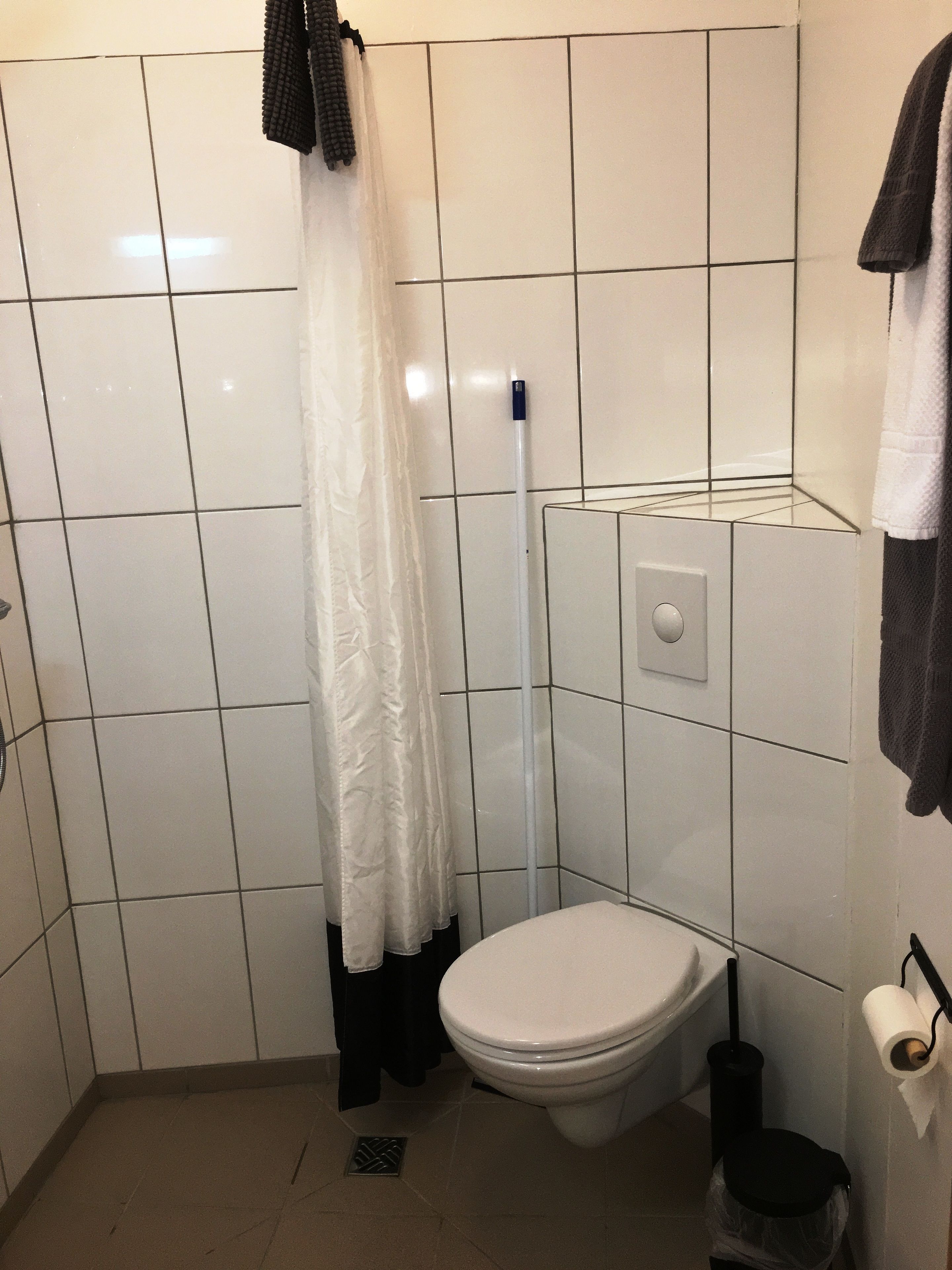A small, tiled bathroom with a toilet, a shower curtain, a mop, and a trash bin.