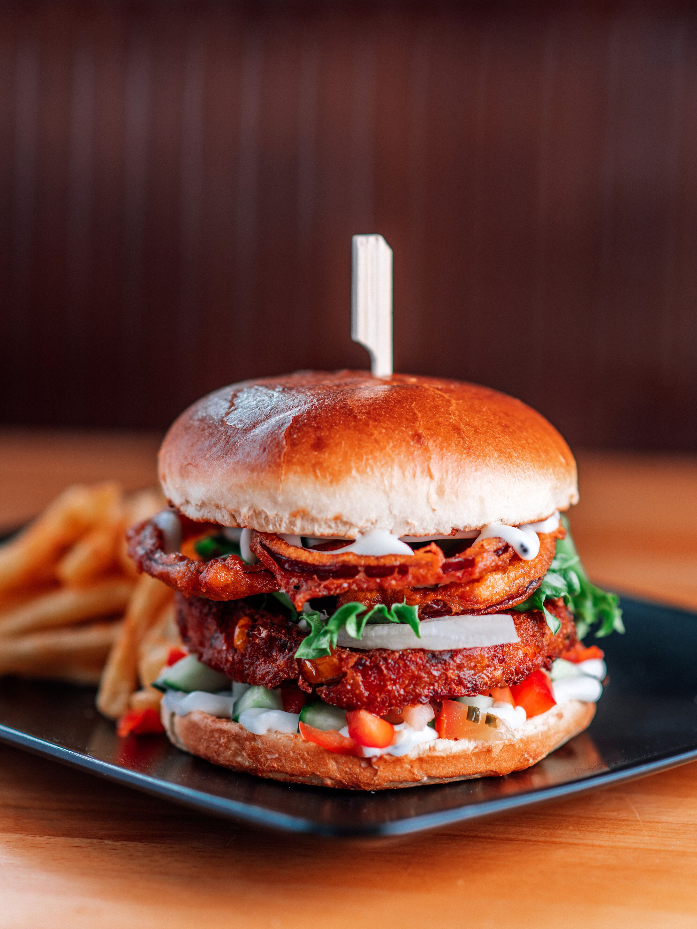 A burger with a toothpick, fries, and a black plate on a wooden table.