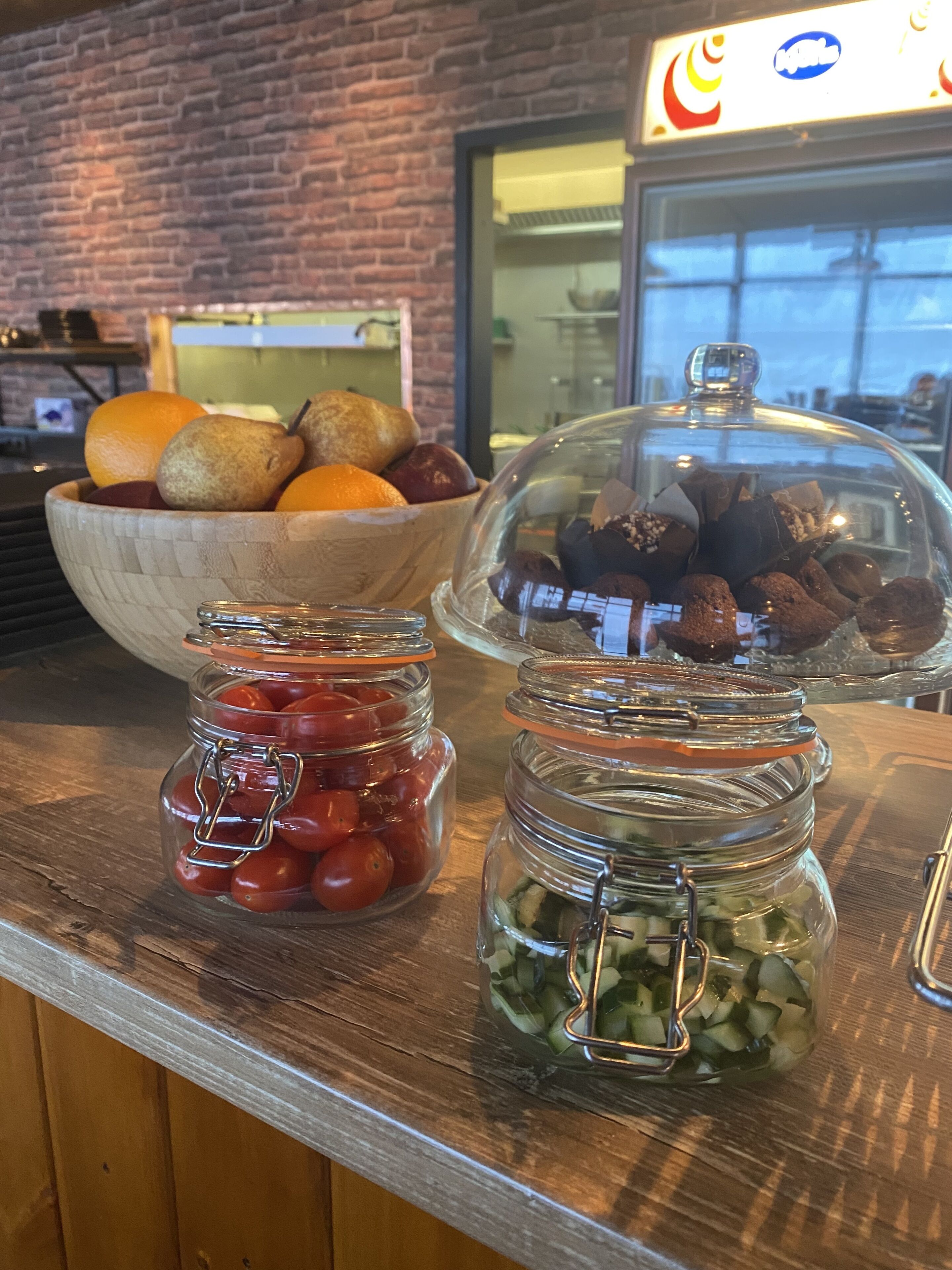 A buffet counter with a wooden bowl of fruits, a glass jar of cherry tomatoes, and a glass jar of sliced cucumbers.