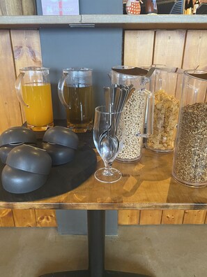 A breakfast bar with various food and drink options, including cereal, a pitcher of orange juice, and a glass of water.