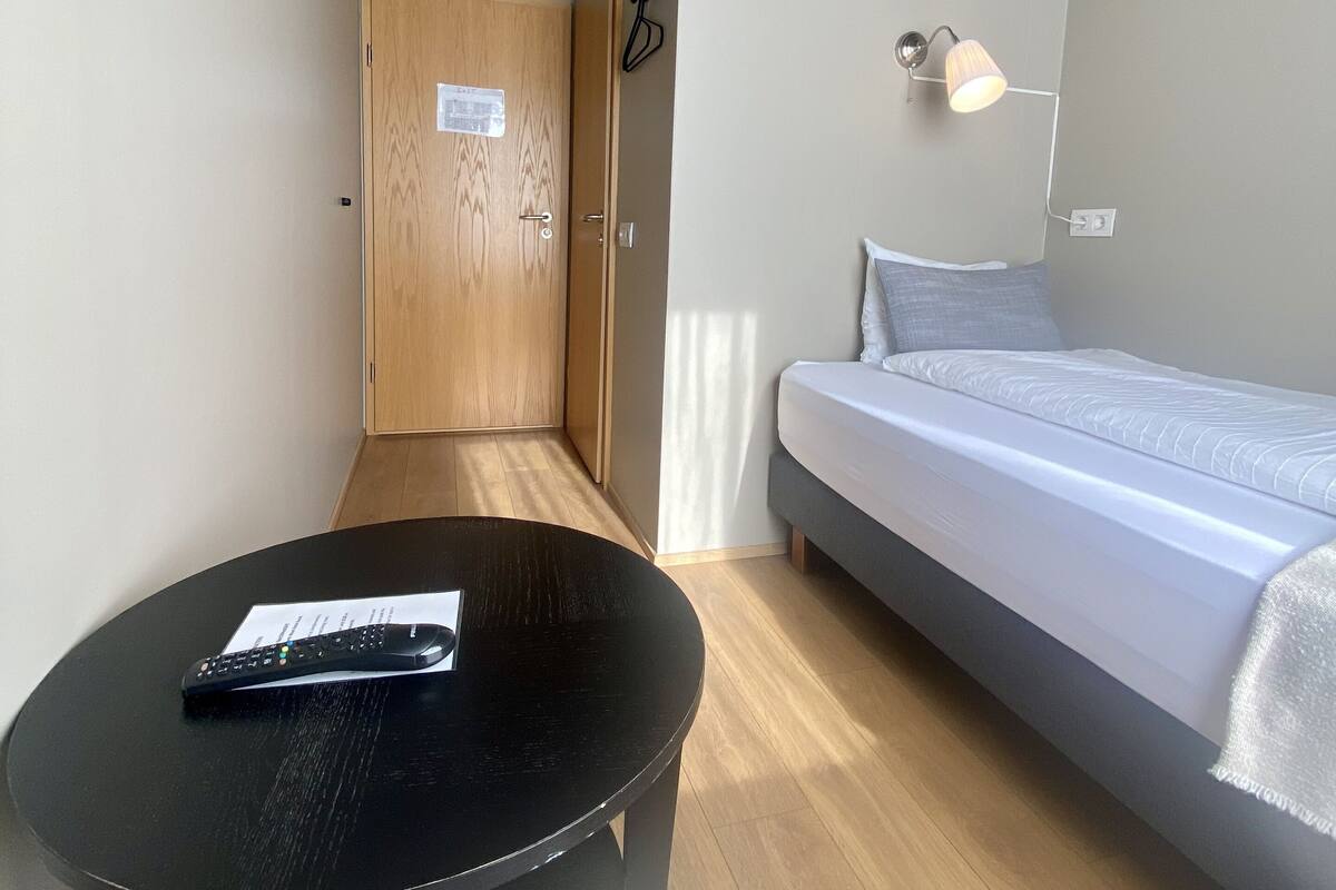 Een moderne hotelkamer met een bed, een ronde zwarte tafel met een afstandsbediening, een klein wit kussen en een houten deur, en een wandlamp.