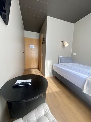 A modern hotel room with a bed, a round black table with a remote control, a small white cushioned seat, a wooden door, and a wall-mounted lamp.