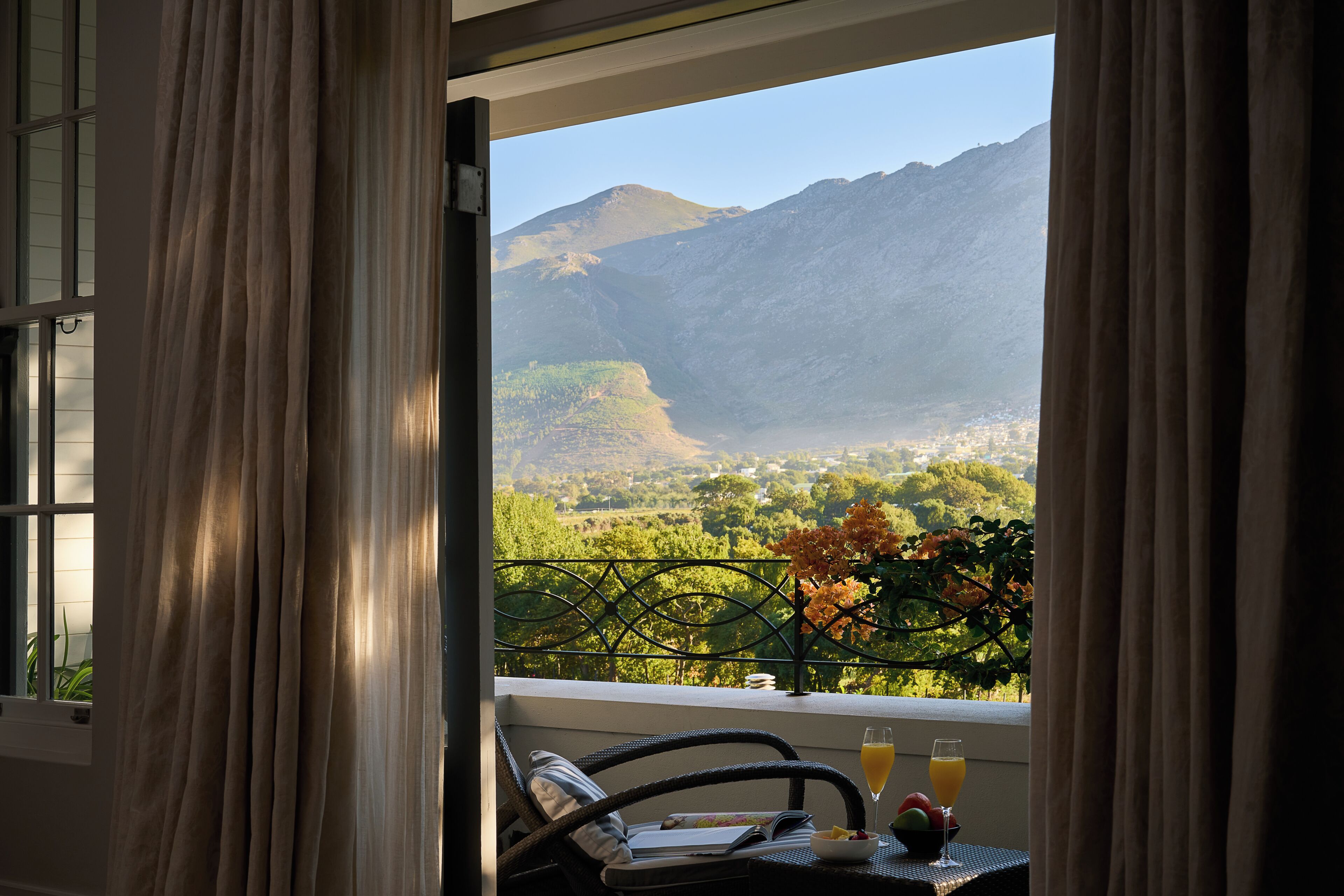 A balcony with a view of mountains, a table with a glass of orange juice, and a chair.