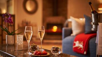 Et veloplyst rum med en pejs, et spisebord med to glas champagne, en vase med blomster og et rødt pled på sofaen.
